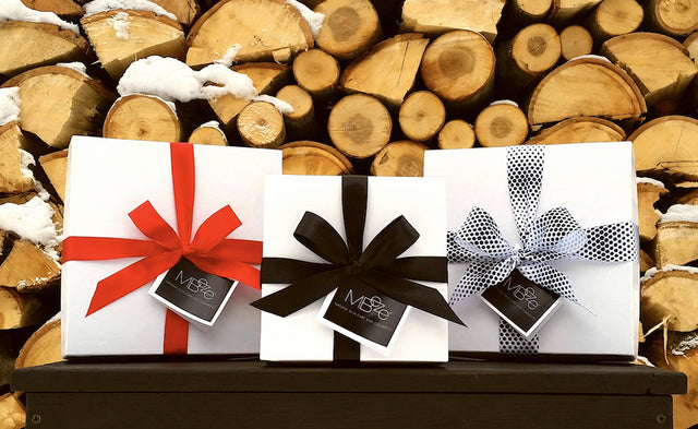 Three MBeze Collection Gift Boxes with stacked, chopped wood behind them and some snow.