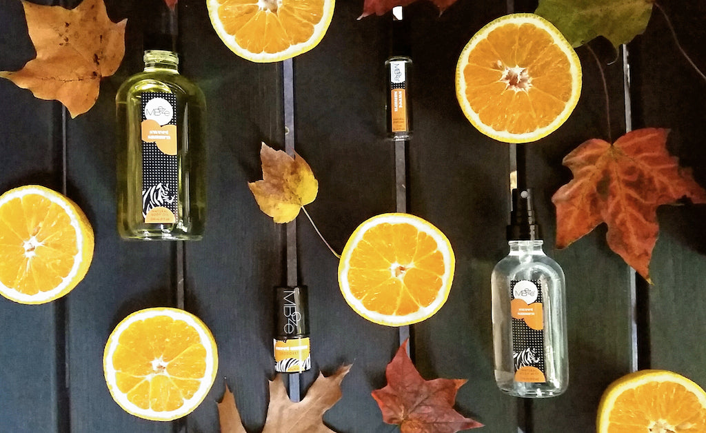 MBeze Sweet Sanura Collection surrounded by oranges sliced in half and fall leaves. Includes the Body Oil, Deodorette, Perfume, and Body Mist.